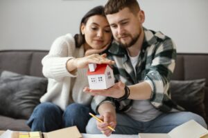 Casal segurando miniatura de casa, sentados em sofá cinza.